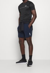Hombre vestido con una camiseta atlética negra, pantalones cortos azul marino con logo lateral, calcetines negros, zapatillas negras y un sudadera negra atada a la cintura.