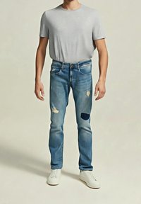Homme portant un t-shirt gris clair, un jean bleu avec des déchirures et des patchs, et des baskets blanches, debout devant un fond uni.