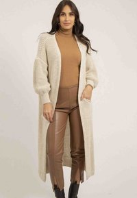 Longue veste en tricot beige à ouverture frontale, manches surdimensionnées et poches, accompagnée de pantalons marron ajustés avec des ourlets fendus.