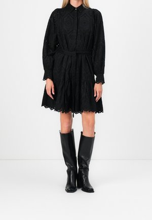 Kvinde iført en sort langærmet skjortekjole med øjesten og bælte samt sorte læderstøvler til knæene, stående foran en hvid baggrund.