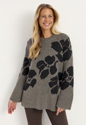 Smilende kvinde med langt blond hår iført en grå sweater med store sorte blomsterprint og mørke bukser mod en ensfarvet baggrund.