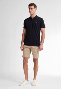 Zwart poloshirt met een klein logo, gecombineerd met beige korte broek met een geometrisch patroon, gedragen met witte sneakers. Simpel, ontspannen pasvorm.
