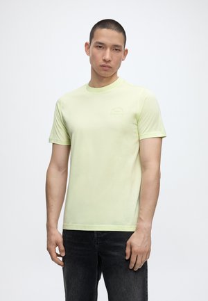 Mladý muž s krátkymi vlasmi, oblečený v bledožltom tričku s krátkym rukávom a čiernych džínsoch, stojaci pred jednoliatym bielym pozadím.