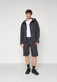 Chaqueta negra resistente al agua con capucha, bolsillo frontal con cremallera, combinada con pantalones cortos oscuros y zapatillas negras. Diseño sencillo y funcional.