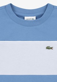 Ανοιχτό μπλε βαμβακερό t-shirt με οριζόντιες λευκές ρίγες, ριμπ λαιμόκοψη και πράσινο λογότυπο κροκοδείλου στην κάτω αριστερή πλευρά.