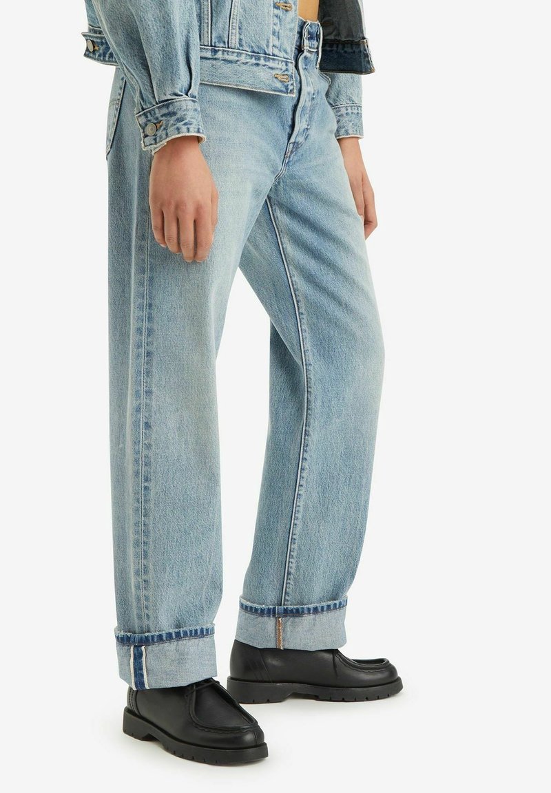 Jean en denim bleu clair, coupe ample, taille haute, ourlets retroussés et coutures contrastées, porté avec des chaussures noires.