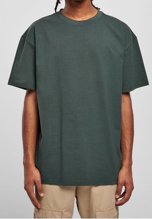 Person iført en mørkegrøn, oversize t-shirt og beige bukser, stående mod en ensfarvet lys baggrund, med armene afslappet ned langs siden.