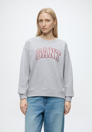 Frau mit langen blonden Haaren trägt ein graues Sweatshirt mit rotem GANT-Logo und hellblaue Jeans vor weißem Hintergrund.