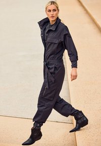 Zwarte jumpsuit met een kraaghals, twee borstzakken, een ceintuur in de taille en taps toelopende broekspijpen, gecombineerd met zwarte puntige laarzen.