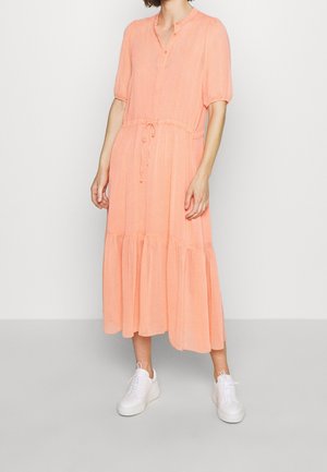 Femme portant une robe midi pêche à manches courtes, une taille à cordon, des boutons au niveau de l'encolure et des baskets blanches, debout sur un sol gris.