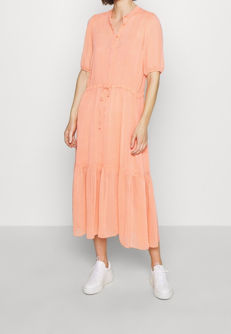 Femme portant une robe midi pêche à manches courtes, une taille à cordon, des boutons au niveau de l'encolure et des baskets blanches, debout sur un sol gris.