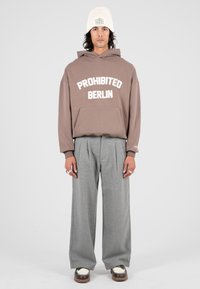 Uomo che indossa una felpa beige con la scritta "PROHIBITED BERLIN", pantaloni grigi a gamba larga, un berretto di maglia bianco e scarpe bicolore, in piedi contro uno sfondo semplice.