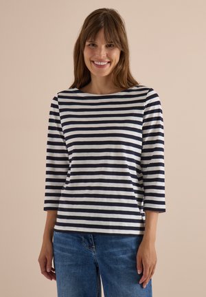 Femme souriante portant un t-shirt à rayures horizontales noires et blanches à manches trois-quarts et un jean bleu sur un fond beige uni.