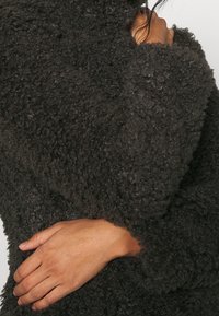 Pull noir et duveteux avec un extérieur texturé et un col haut ; présente des manches ajustées et amples et un aspect doux et confortable.