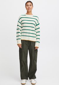 Maglione a righe con strisce orizzontali verdi e crema, texture a costine, abbinato a pantaloni cargo verdi oliva larghi e sneakers bianche.