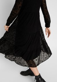 Person som bär en svävande svart spetsklänning med långa ärmar och svarta snörstövlar, mitt i ett steg mot en enkel bakgrund.