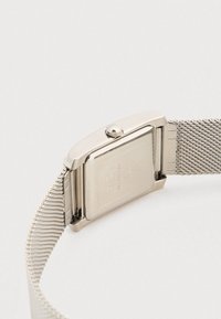 Ασημένιο ορθογώνιο ρολόι χειρός με κορδέλα από μεταλλικό πλέγμα απλωμένο επίπεδα, που δείχνει το χαραγμένο πίσω κάλυμμα και το κορώνα ρύθμισης σε λευκή επιφάνεια.