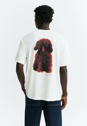 Persona con capelli corti e ricci che indossa una maglietta bianca con una grande stampa di un cane soffice rosso e nero sulla schiena, abbinata a jeans blu scuro.