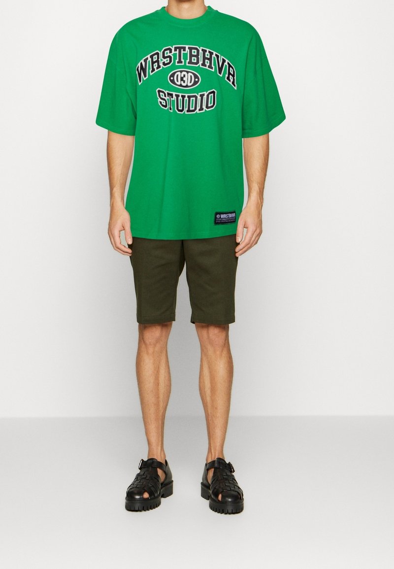 Groene oversized t-shirt met zwarte tekst "WRSTBHVR STUDIO", gecombineerd met olijfgroene shorts en zwarte sandalen. Eenvoudige, casual stijl.