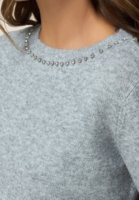 Gros plan sur une personne portant un pull en maille gris clair avec un col rond orné d'une rangée de petits strass transparents.