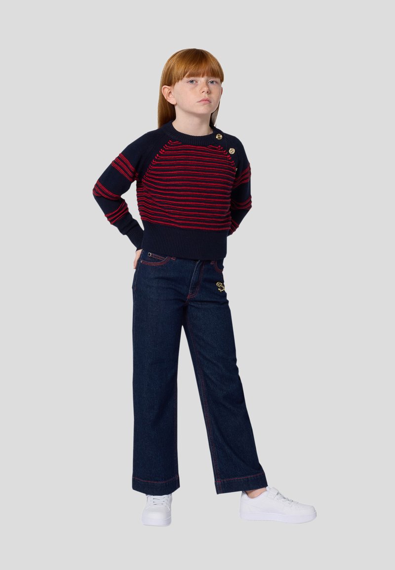 Marineblauwe trui met rode strepen en knoopaccenten, gecombineerd met donkere spijkerbroeken met wijde pijpen en witte sneakers. Soepele textuur en een ontspannen pasvorm.