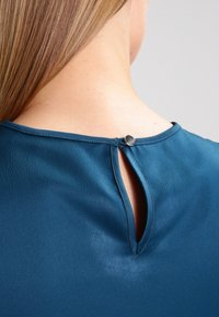 Turkusowa bluzka z okrągłym dekoltem i wycięciem w kształcie łezki z tyłu, zapinaną na mały srebrny guzik, uszyta z gładkiego materiału.