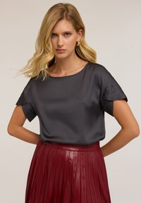 Blusa in satin color antracite con maniche corte e strutturate, abbinata a una gonna plissettata bordeaux. Tessuto liscio e vestibilità rilassata.