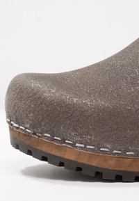Gros plan sur le bout d'une chaussure marron texturée avec des coutures visibles, une semelle intermédiaire en bois et une semelle extérieure en caoutchouc noir crantée.