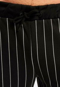 Pantaloni de trening negri din bumbac cu dungi verticale albe, având un brâu elastic, șnur ajustabil și o textură netedă.