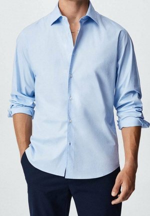 Mies, jolla on päällään vaaleansininen pitkähihainen kauluspaita, hihat käännettynä ja tummat housut, seisoo yhdellä kädellä taskussa.