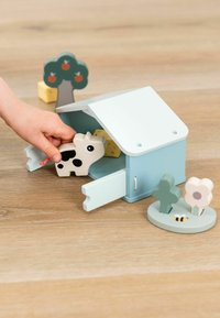 Houten speelset met een lichtblauw schuur, openingen, een koefiguur, bloemen- en boomaccessoires, en een natuurlijk houten oppervlak.