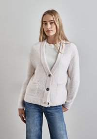 Cardigan di colore crema chiaro in morbido tessuto lavorato, con polsini a coste, grandi bottoni frontali e due tasche laterali, abbinato a jeans blu.