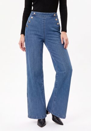 Jean bleu taille haute à jambes larges avec des boutons dorés sur les hanches, porté avec un haut noir à manches longues côtelé et des bottes noires à bout pointu.