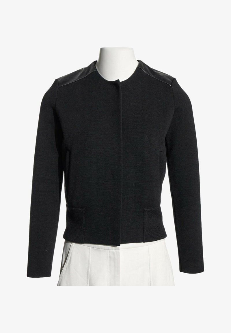 Veste noire à manches longues avec des panneaux en cuir aux épaules et sans boutons visibles, présentée sur un buste de mannequin blanc portant un pantalon blanc.