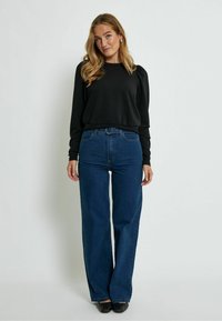 Top nero a maniche lunghe con spalle a sbuffo, abbinato a jeans blu a vita alta e scarpe nere flat, su uno sfondo bianco semplice.