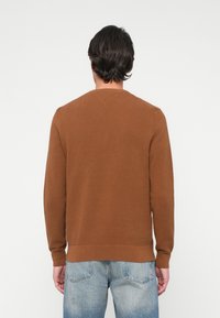 Brauner strukturierter Pullover mit langen Ärmeln und Rundhalsausschnitt, kombiniert mit hellblauen Jeans. Der Stoff hat ein dezentes gewebtes Muster.