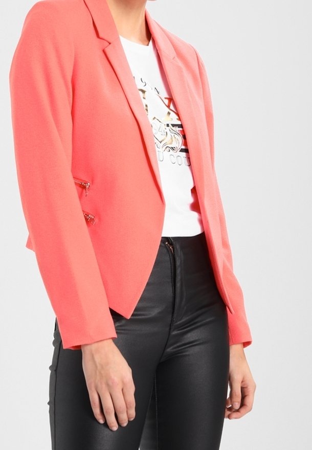 Blazer corail au design structuré, avec deux accents zippés à l'avant, porté sur un t-shirt blanc à motif graphique, associé à un pantalon en cuir noir.