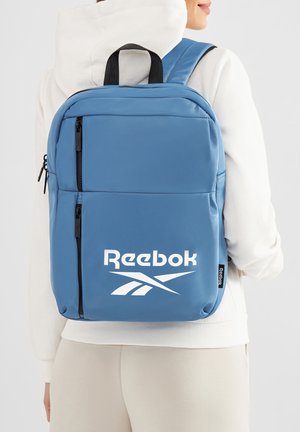 Persona con una mochila azul de Reebok con cremalleras negras sobre una sudadera blanca con capucha, vista desde atrás y de lado.
