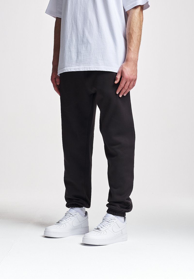 Schwarze Jogginghosen mit elastischen Bündchen, kombiniert mit weißen Sneakers und einem lockeren weißen T-Shirt, das einen lässigen, entspannten Stil unterstreicht.