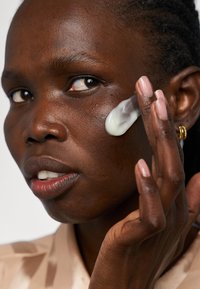 Une crème hydratante est appliquée sur la joue d'une personne à la peau foncée. La crème est blanche et la personne a les cheveux courts et tressés.