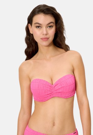 Mujer joven con cabello largo y ondulado de color castaño, vistiendo un top de bikini rosa texturizado sin tirantes, sobre un fondo liso.