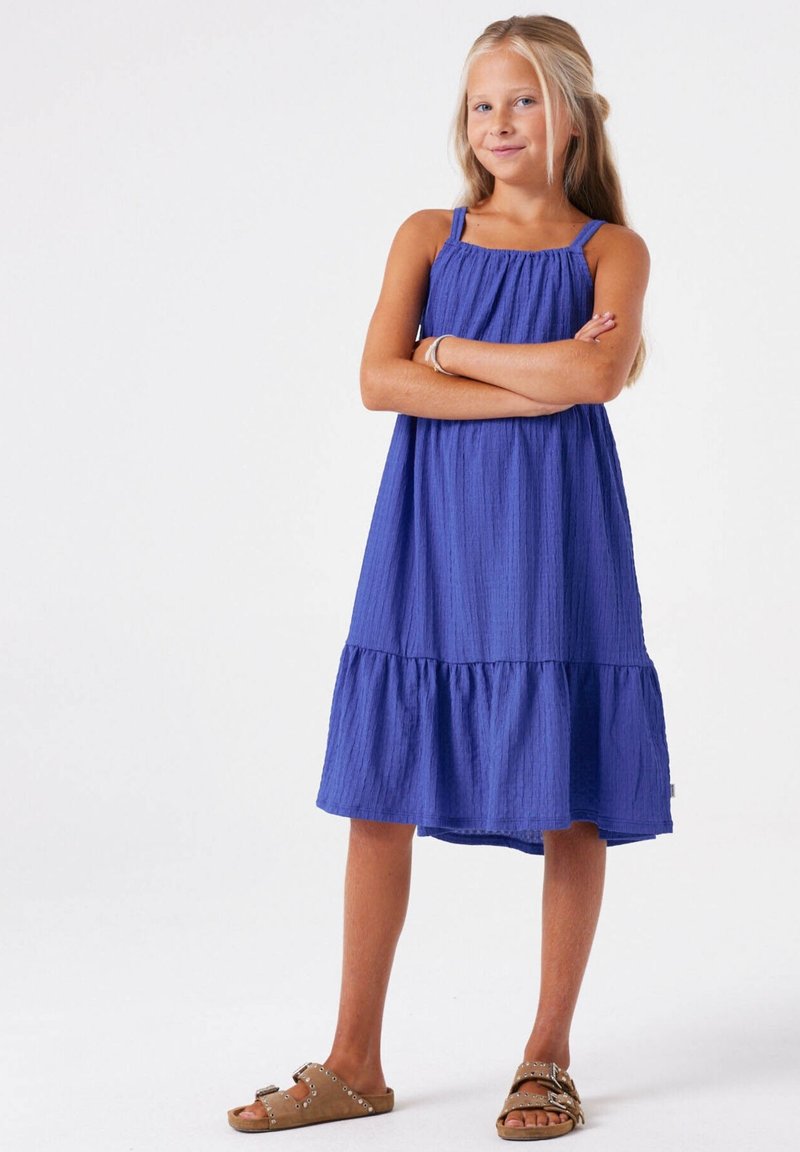 Robe bleu royal à volants avec fines bretelles et tissu texturé, dotée d'un ourlet à volants. Associée à des sandales marron.