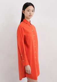 Chemise rouge à manches longues, en tissu lisse. Avec un col pointu et un léger détail de boutons sur le devant.
