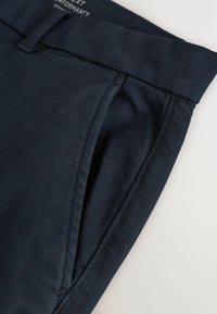 Granatowe spodnie o gładkiej fakturze, z bocznymi kieszeniami, płaskim przodem i tkanym pasem. Płaskie szwy widoczne wzdłuż szwów.