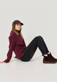 Burgundy knit sweater, black jeans, and dark ankle boots with textured soles. Styled with a black cap, the model sits against a neutral background.