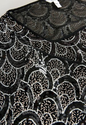 Schwarz schimmernder Stoff mit einem Muschel-Muster, das silberne und beiges Akzente aufweist. Weiche Textur mit glänzenden, reflektierenden Oberflächen.