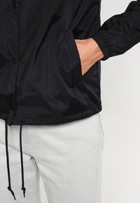 Czarna nylonowa kurtka z zatrzaskami, elastycznymi mankietami i dwiema bocznymi kieszeniami. Dolna krawędź ze sznurkiem. Nosi się ją na jasnych dżinsowych spodniach.