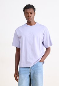 Lilla kortærmet t-shirt med rund hals og et lille logo på brystet. Kombination med lyseblå denimjeans. Ensfarvet baggrund.