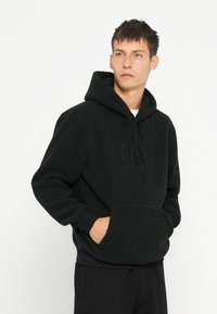 Schwarzer Fleece-Hoodie mit einer Kängurutasche, strukturiertem Stoff, Kordelzug-Kapuze und gerippten Bündchen. Mit gesticktem Logo auf der Vorderseite.