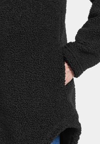 Sort fleecejakke med en tekstureret overflade, sidelomme og en kant, der taper til en spids. Ærmerne har en løs pasform.